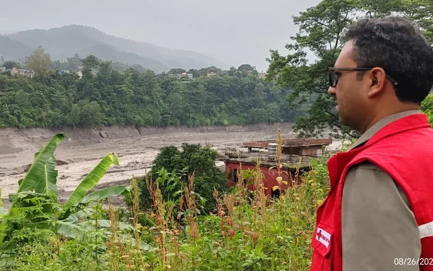 A Nepal Red Cross volunteer in a red vest looks out over a flooded river valley