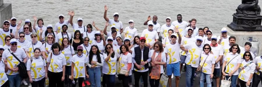 A large group of Rudy A Menon Foundation supporters in matching white and yellow T-shirts wave beside a river