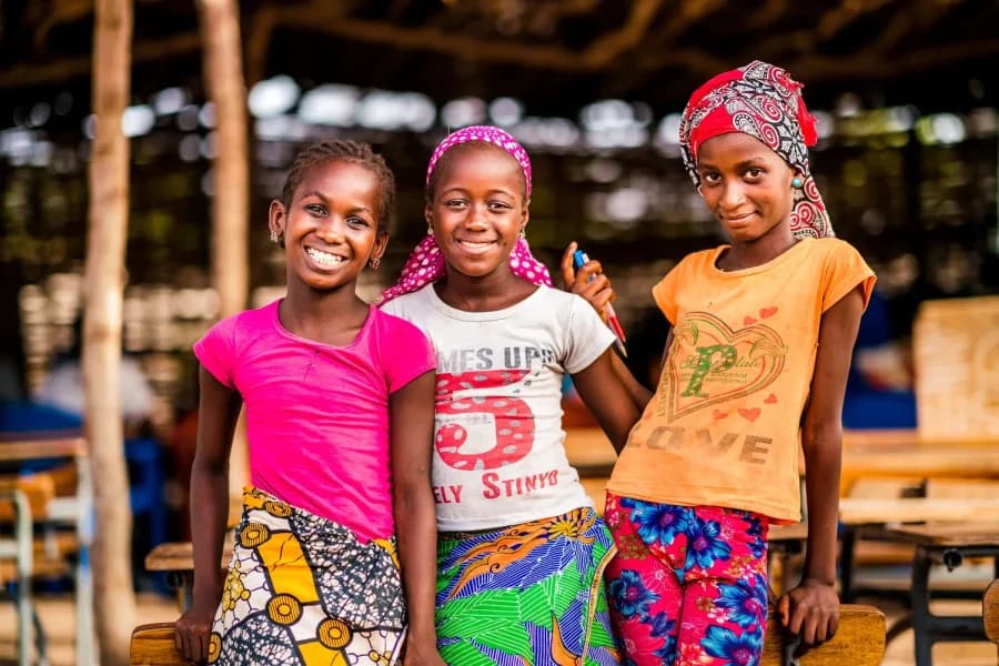 Three smiling girls in bright dresses stand together, supported by Save the Children